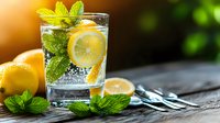 Refreshing glass of sparkling water with lemon slice, fresh mint leaves and ice cubes on rustic wooden table, citrus fruits nearby for summer drink.