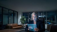 Mature woman with gray hair and light complexion, wearing professional business attire, holding tablet with glowing digital interface in modern office environment at night.