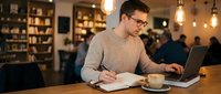 Young man with glasses, Caucasian ethnicity, wearing beige sweater, working on laptop while taking notes in notebook at wooden table with coffee cup in modern library or coworking space.