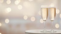 Two elegant champagne flutes filled with sparkling wine against soft bokeh background in beige and silver tones. Romantic celebration setting with blurred lights.