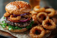 Gourmet beef burger with crispy onion rings, lettuce, red onion slices and bacon on sesame bun served with golden fried onion rings on wooden board.