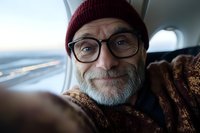Cheerful mature Caucasian man with gray beard wearing glasses, red beanie and brown sweater taking selfie while traveling by airplane, genuine smile.