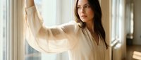 Young brunette woman in flowing white blouse standing by window with natural light, peaceful morning moment at home for wellness and lifestyle content.