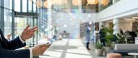 Businessman in dark suit gestures toward digital network interface with glowing nodes and connecting lines in modern office lobby with glass windows and colleagues in background.
