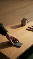 Hand reaching for a smartphone on a wooden desk with a ceramic mug and pencils, warm moody lighting, modern workspace concept.