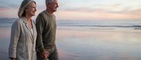 Happy senior couple holding hands walking on beach at sunset enjoying romantic vacation together during golden hour with ocean waves.