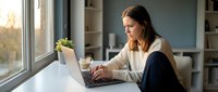 Young professional woman working on laptop computer at modern home office desk near window with natural sunlight and plants.