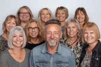 Mature man with gray beard surrounded by group of smiling middle-aged women with varied hairstyles against neutral gray background, representing friendship or team portrait.
