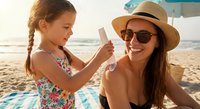 Young girl applying sunscreen to smiling woman's face on sunny beach. Mother and daughter enjoying summer vacation with sun protection skincare routine.