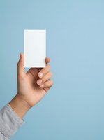 Hand of a person with medium skin tone holding a blank white card against a light blue background for branding and marketing mockups.