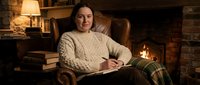 Young woman in cozy cream sweater reading book by fireplace in rustic living room with warm lighting and comfortable armchair for relaxation content.