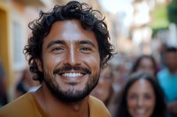 Hispanic man with curly dark hair and bright white smile wearing yellow t-shirt looking at camera with genuine happiness, crowd in background on urban street.