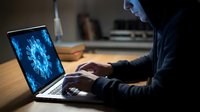 Hooded hacker working on laptop with virus malware code on screen in dark room, cybersecurity threat concept for digital security awareness campaigns.