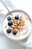 Creamy yogurt bowl topped with fresh blueberries, pine nuts, almond butter and cinnamon powder in white ceramic bowl on light background. Food photography.