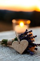 Romantic composition with dried lavender bouquet and decorative burlap heart on lace fabric, glowing candle creating warm atmosphere at sunset.