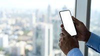 Male professional with dark skin tone holding smartphone with blank white screen against urban cityscape window view from high-rise office building.