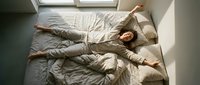 Young woman with long brown hair, lying on unmade bed in casual beige clothing, arms stretched overhead in relaxed pose, natural daylight from window, overhead view.
