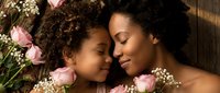 Mother and daughter with curly hair embrace surrounded by pink flowers and baby's breath, sharing a tender moment together on wooden background.