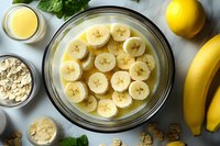 Fresh banana slices soaking in glass bowl with liquid, surrounded by whole bananas, oats, mint leaves and lemons on light gray surface for healthy breakfast.