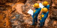 Construction workers in blue uniforms and yellow hard hats inspect excavation site, reviewing documents while standing on orange clay soil at mining or building location.