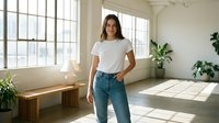 Young Hispanic woman in casual white t-shirt and jeans standing confidently in bright modern apartment with large windows and green plants.