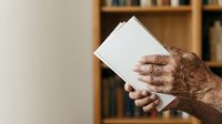 Senior man with age spots holding blank white tablet device in home library setting with wooden bookshelf background. Suitable for technology adoption, elderly users, digital literacy.