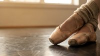 Close-up of ballet dancer's feet in worn pointe shoes on wooden studio floor with warm natural light streaming through windows creating artistic shadows.