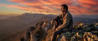 Contemplative man sitting on mountain peak during golden hour sunset with dramatic orange sky and flying birds in distance.