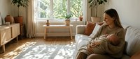 Young Caucasian mother sitting on sofa breastfeeding newborn baby in bright Scandinavian living room with natural light and plants.