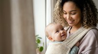 Young mixed-race mother with curly hair holding smiling baby in carrier wrap at home window, bonding moment for parenting and childcare content.
