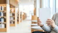 Person with light skin tone holding blank white book in library setting with wooden shelves and natural light, suitable for education and publishing concepts.