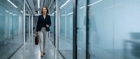 Professional businesswoman walking confidently through modern glass office corridor with briefcase, corporate executive in formal attire.