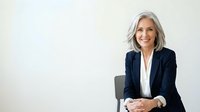 Mature woman with gray hair and fair complexion wearing dark navy blazer and white shirt, seated on gray chair against white background, smiling at camera with hands clasped, suitable for corporate and business portraits.