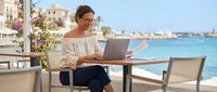 Professional middle-aged Caucasian woman working remotely on laptop at seaside cafe terrace with Mediterranean coastal view, digital nomad lifestyle concept.
