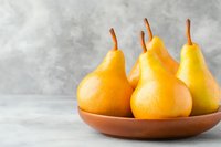 Fresh ripe yellow pears arranged in ceramic bowl on light gray background, minimal food composition with natural lighting and soft shadows.