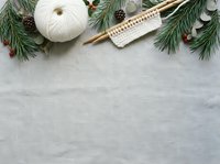 Flat lay composition with evergreen branches, cream yarn ball, and wooden knitting needles on gray linen background, ideal for craft and holiday projects.