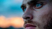 Close-up portrait of young Caucasian man with striking blue eyes and beard against blurred sunset background, conveying determination and contemplation.