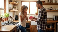 Young African American man surprising his girlfriend with gift box in cozy kitchen while she covers face with hands in excitement and joy.