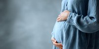 Pregnant woman in cozy blue sweater gently holding belly with both hands against soft gray background for maternity and motherhood concepts.