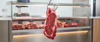 Raw beef steak hanging on hook in front of butcher shop display case with fresh meat selection, ready for sale or preparation in professional butchery environment.