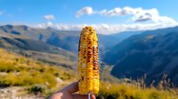 Grilled corn on cob held up against scenic mountain landscape background with blue sky and white clouds, showcasing outdoor dining and adventure lifestyle.