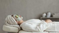 Young Caucasian woman relaxing at spa with clay face mask and cucumber slices, lying on treatment table in white robe and towel turban.