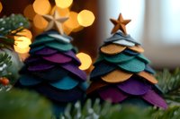 Handcrafted felt Christmas trees in vibrant teal, purple, and gold tones with star toppers against blurred holiday lights and evergreen branches create festive decor.