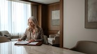 Professional mature Caucasian businesswoman in beige suit working with documents at modern office desk near window with natural light and contemporary decor.