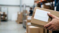 Worker holding cardboard box with blank label in warehouse storage facility with shelves and packages in background for shipping and logistics.