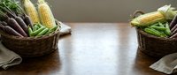 Wicker baskets filled with fresh corn and purple beans on wooden table, ideal for farmers market, grocery, and food styling projects.