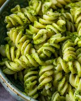 Rotini pasta with fresh pesto sauce and herbs in ceramic bowl, macro food photography showing texture and spiral shapes of cooked pasta noodles.