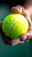 Close-up macro shot of bright yellow tennis ball held in fingers against dark green background, showing detailed texture and white curved line pattern.