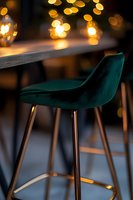 Elegant emerald velvet bar stool with gold metal legs against blurred bokeh lights background creates luxurious atmosphere in modern restaurant interior.