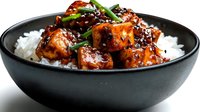 Glazed teriyaki chicken pieces with sesame seeds served over steamed white rice in black bowl, garnished with fresh green onions for Asian cuisine meal.